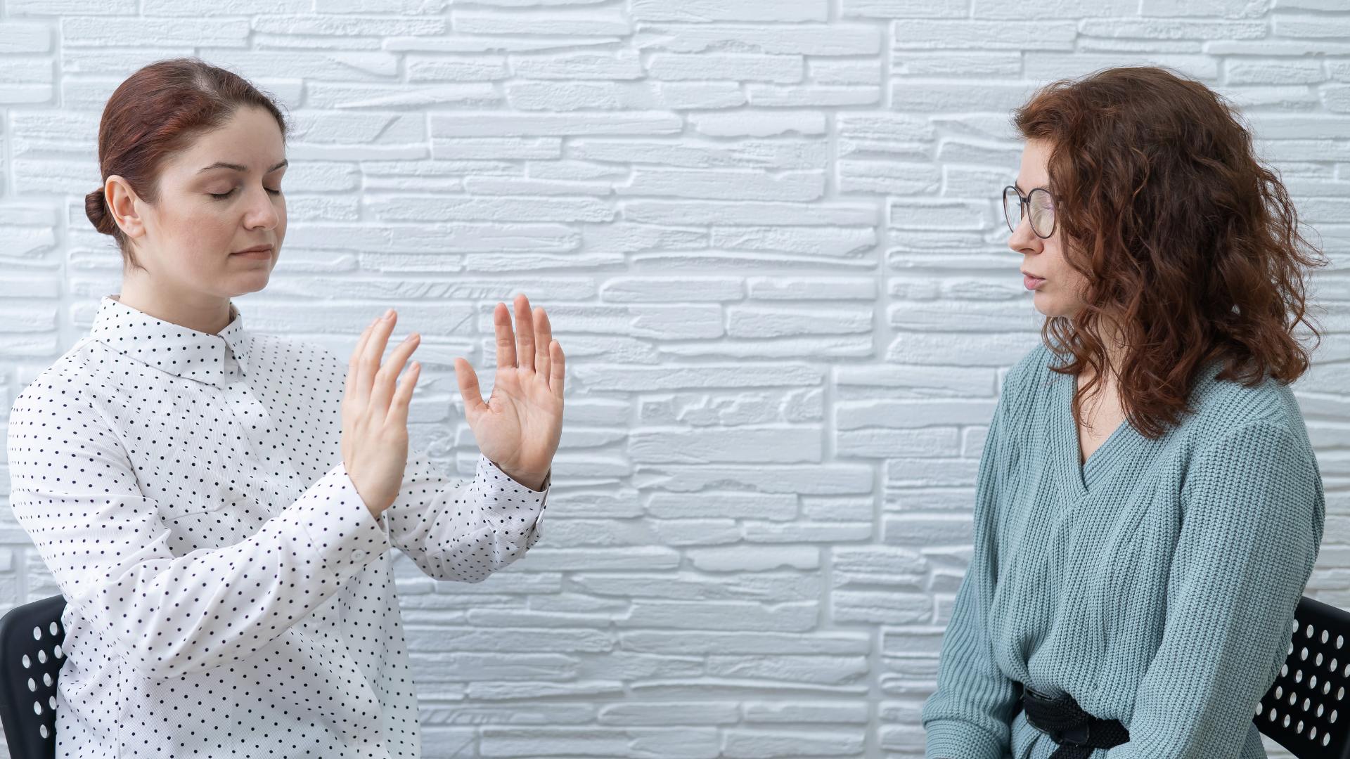 Photo of two woman, one woman is guiding the other into a hypnotic trance, the woman being guided has her hands raised whilst the rest of her relaxes with her eyes closed.
