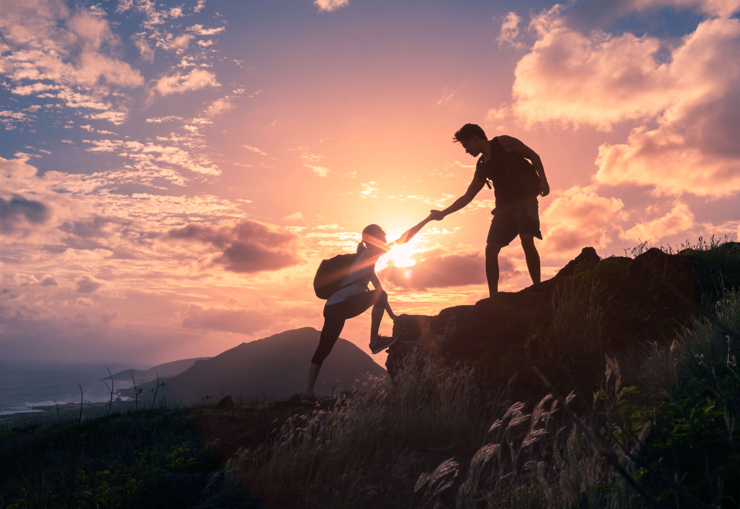 A photo of two people helping each other hike up a mountain at sunrise.